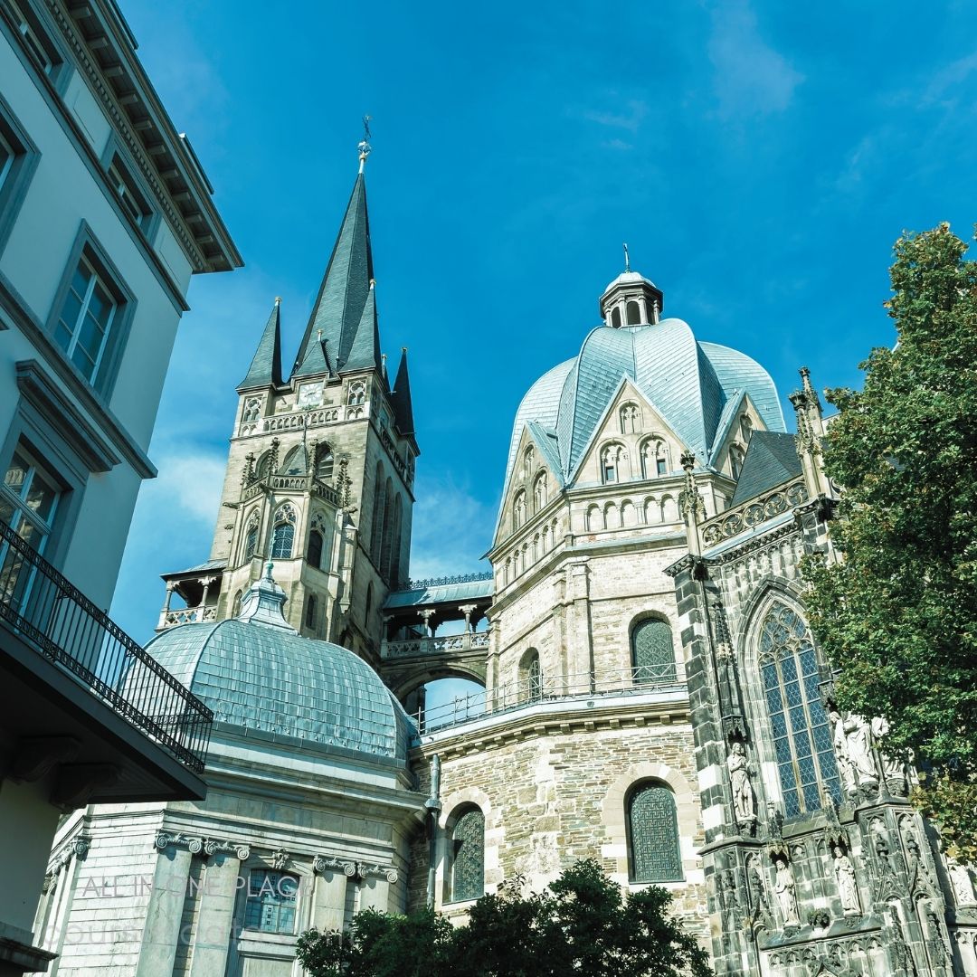 Historic cathedral with domes. Tall spire touching the sky. Intricate architectural details visible. Surrounded by greenery and buildings. Clear blue sky backdrop.