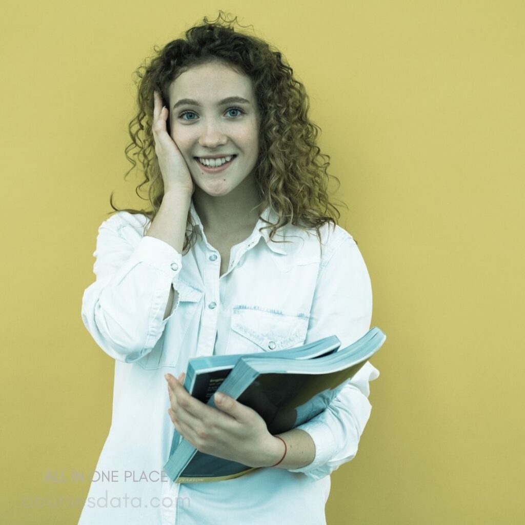 Smiling young woman, curly hair. White shirt, holding books. Bright yellow background. Positive and enthusiastic expression.