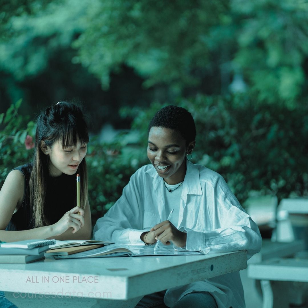 Two students studying together. Smiling young women. Outdoor setting with greenery. Notebook and pencil on table.