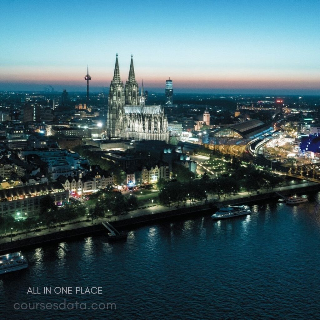 Cologne Cathedral at dusk. City skyline with lights. River reflecting city lights. Trains at station foreground. Colorful buildings along waterfront.