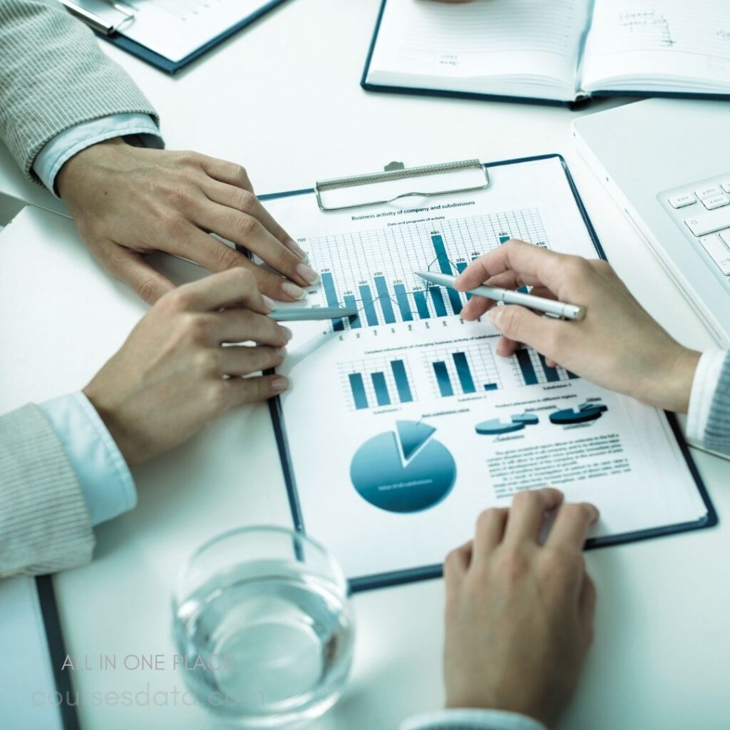 Hands analyzing business charts. Graphs and data visualization. Documents on a conference table. Clear glass of water nearby. Office setting with laptops.