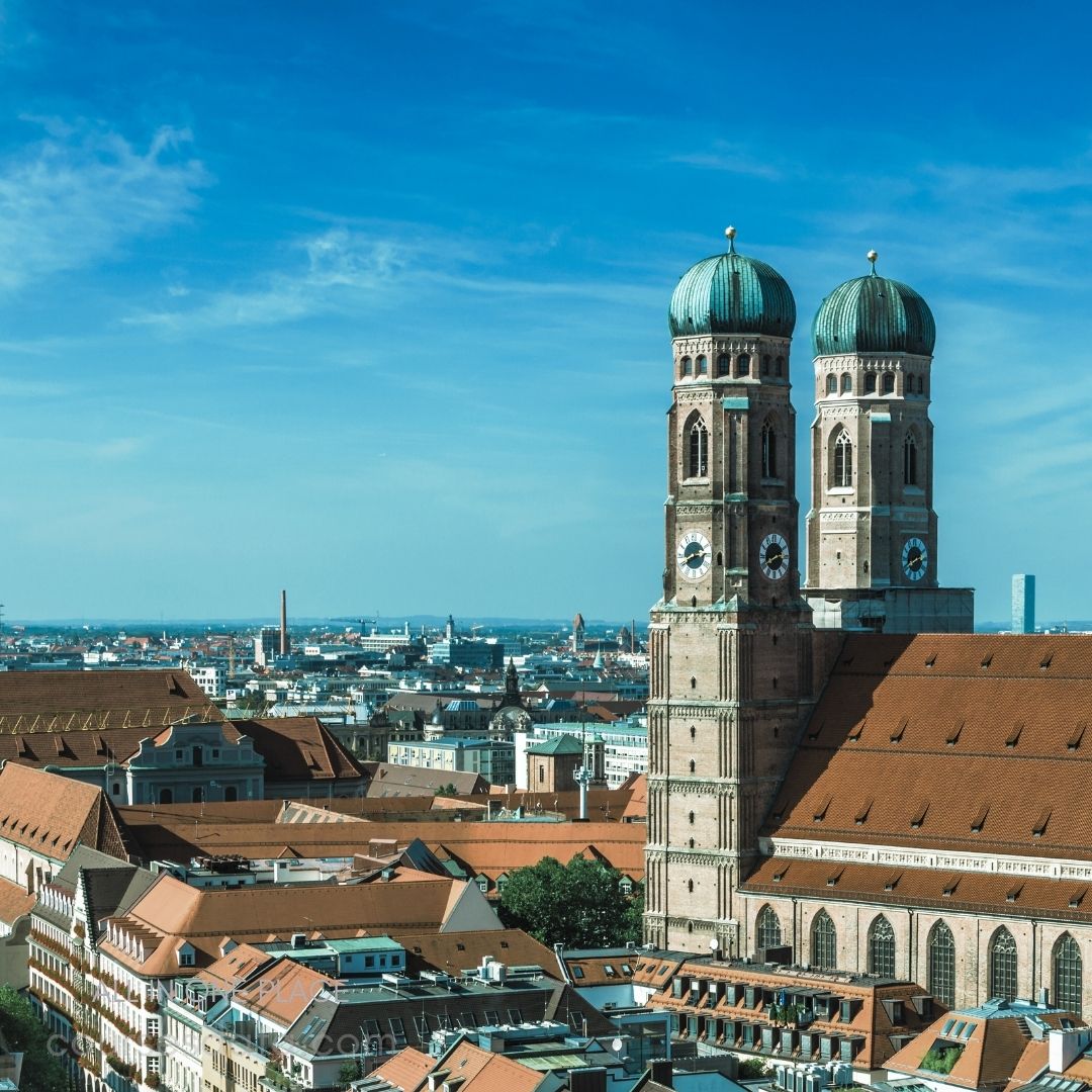 Munich skyline with church towers. Blue sky and city rooftops. Distinctive green-domed towers visible. Historic architecture in foreground. Vast cityscape stretches into distance.