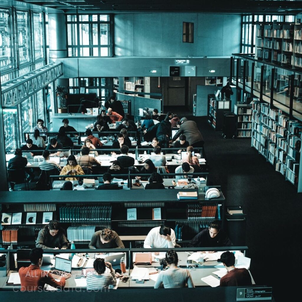 Busy library study environment. Students engaged in studying. Tables filled with books. Natural light through large windows. Bookshelves lining the walls.