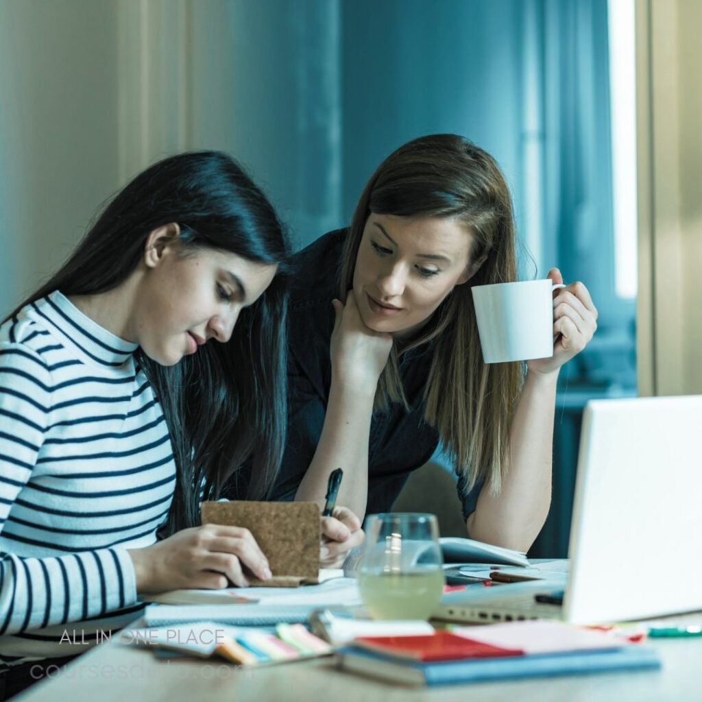 Two women collaborating at table. One writes in notebook. The other holds coffee cup. Laptop and stationery visible. Bright, modern workspace atmosphere.