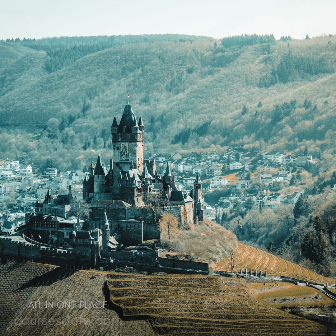 Castle on a hillside. Surrounded by green mountains. Nearby picturesque village. Vineyards in the foreground. Clear blue sky.
