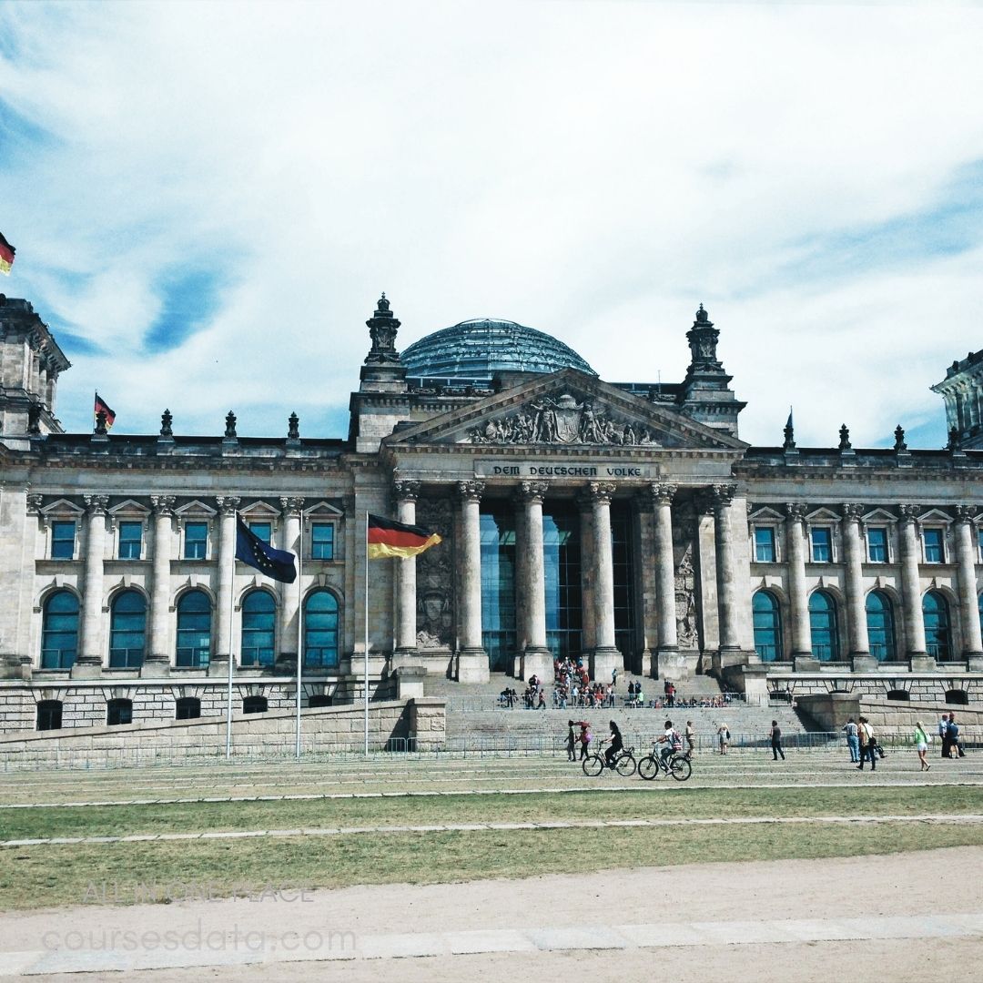 Reichstag building, Berlin. European and German flags. Visitors and cyclists present. Cloudy blue skies above. Historic architecture and steps.