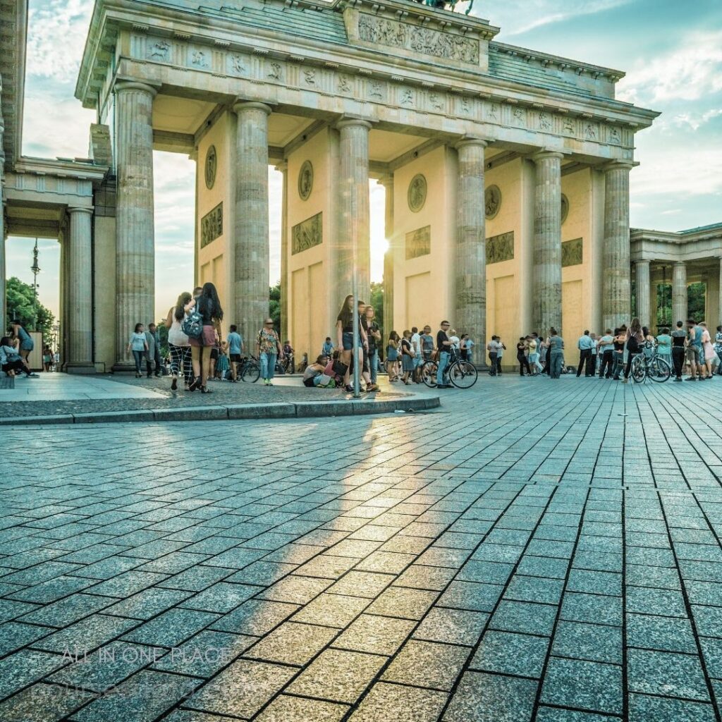 Brandenburg Gate in Berlin. Crowd of people gathering. Shimmering reflective pavement. Golden hour sunset lighting. Bicycles parked nearby.