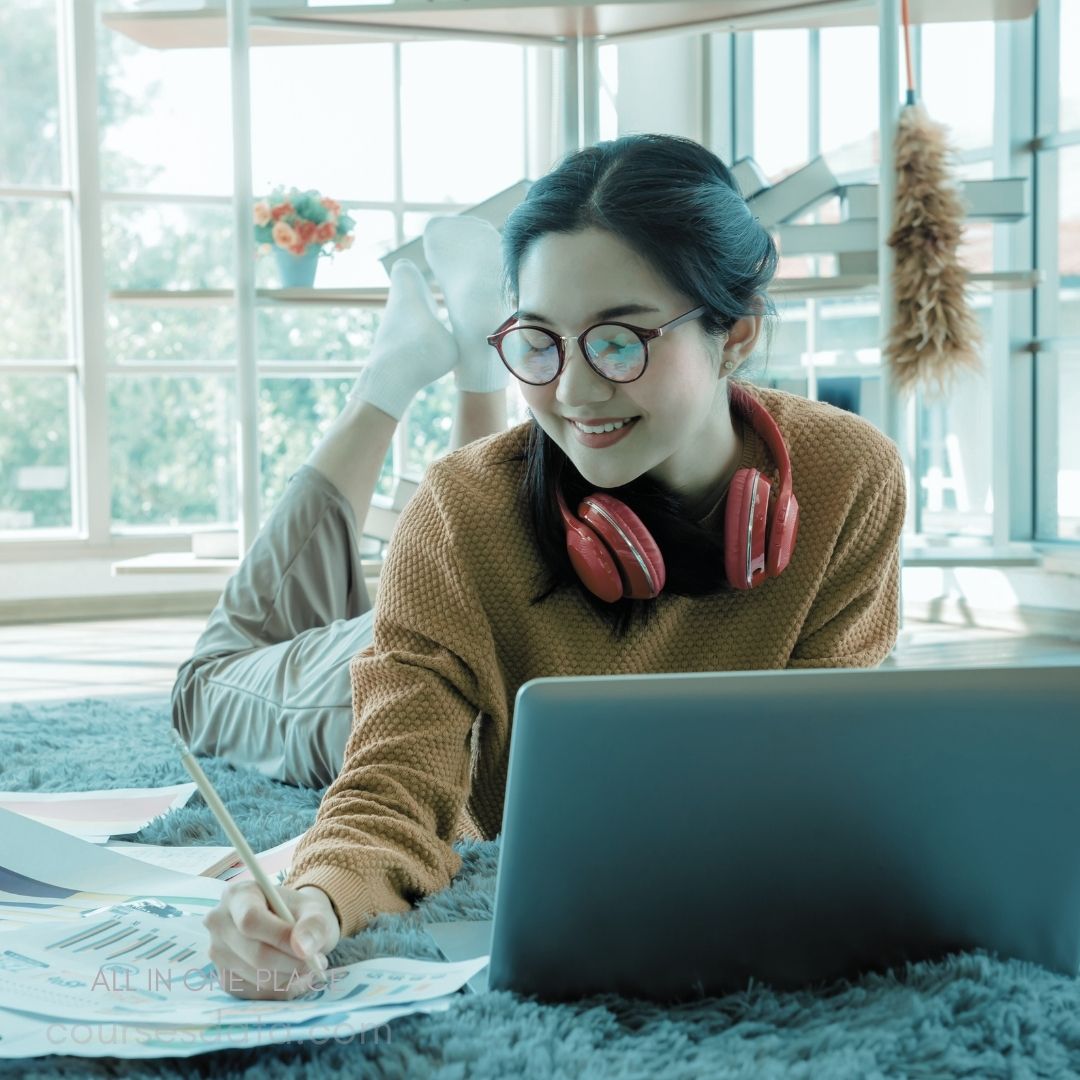 Young woman studying at home. Wearing red headphones and glasses. Lying on a cozy carpet. Writing notes with a pencil. Laptop and documents scattered nearby.