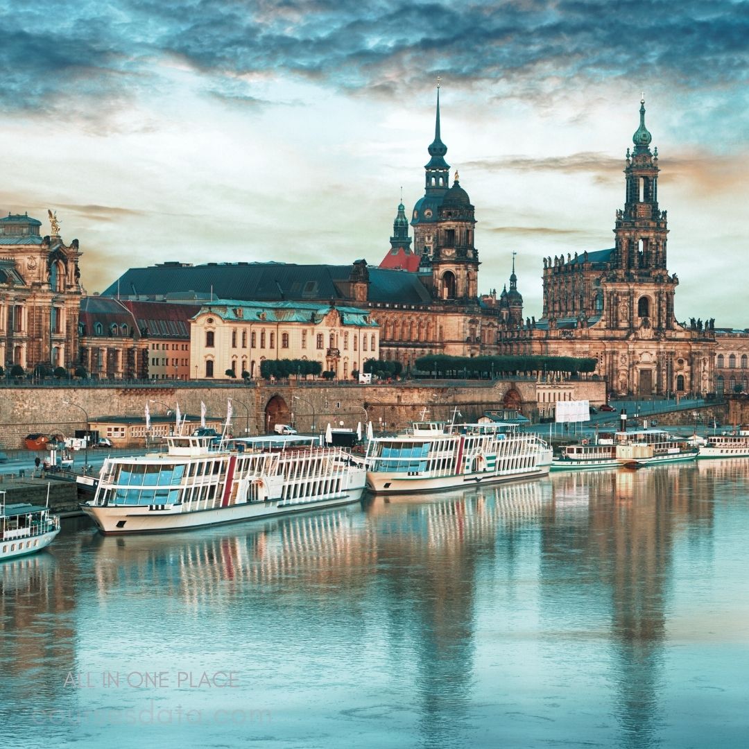 Historic buildings along riverbank. Boats docked on tranquil water. Dramatic sky with clouds. Spire towers above architecture. Peaceful scene at dusk.