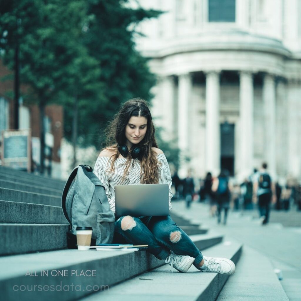 Young woman studying outdoors. Sitting on steps, laptop open. Wearing headphones, casual attire. Coffee cup and backpack nearby. Historical building in background.