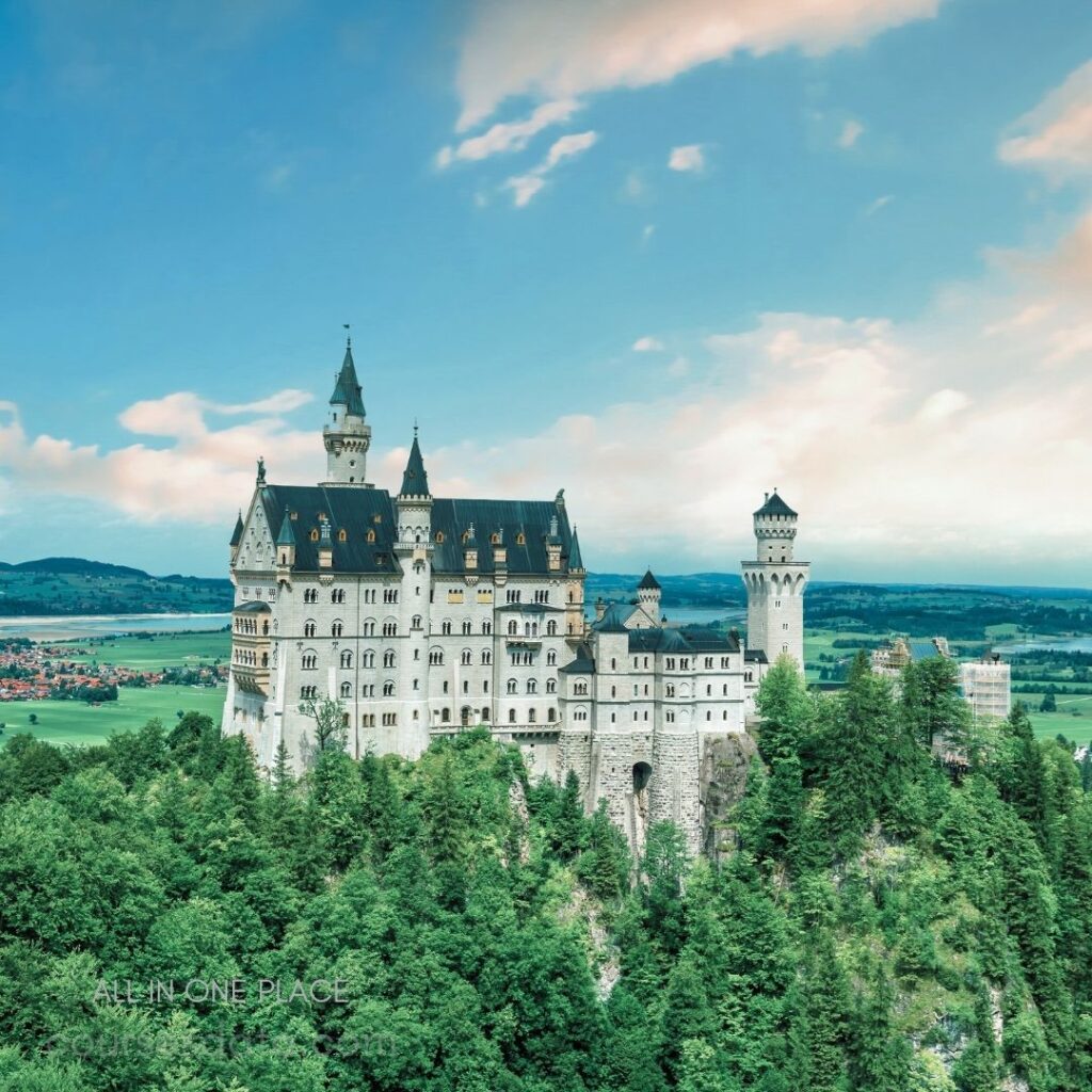 Fairy-tale castle on hilltop. Surrounded by lush green trees. Bright blue sky in background. Historic architecture with towers. Scenic landscape and distant mountains.