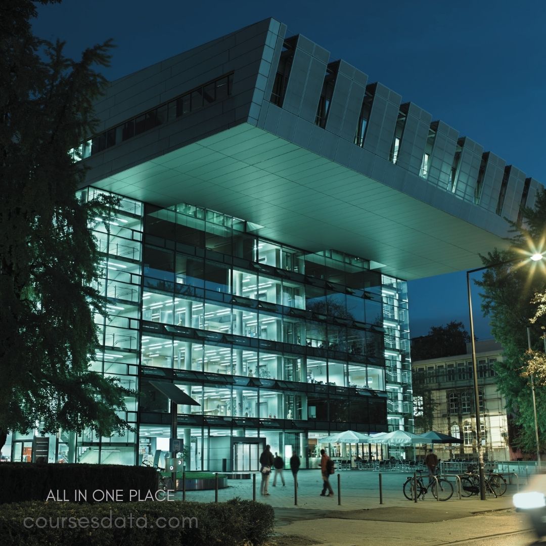 Modern glass building at night. Illuminated interior spaces visible. People walking in foreground. Surrounding trees and streetlights. Café seating area outside.