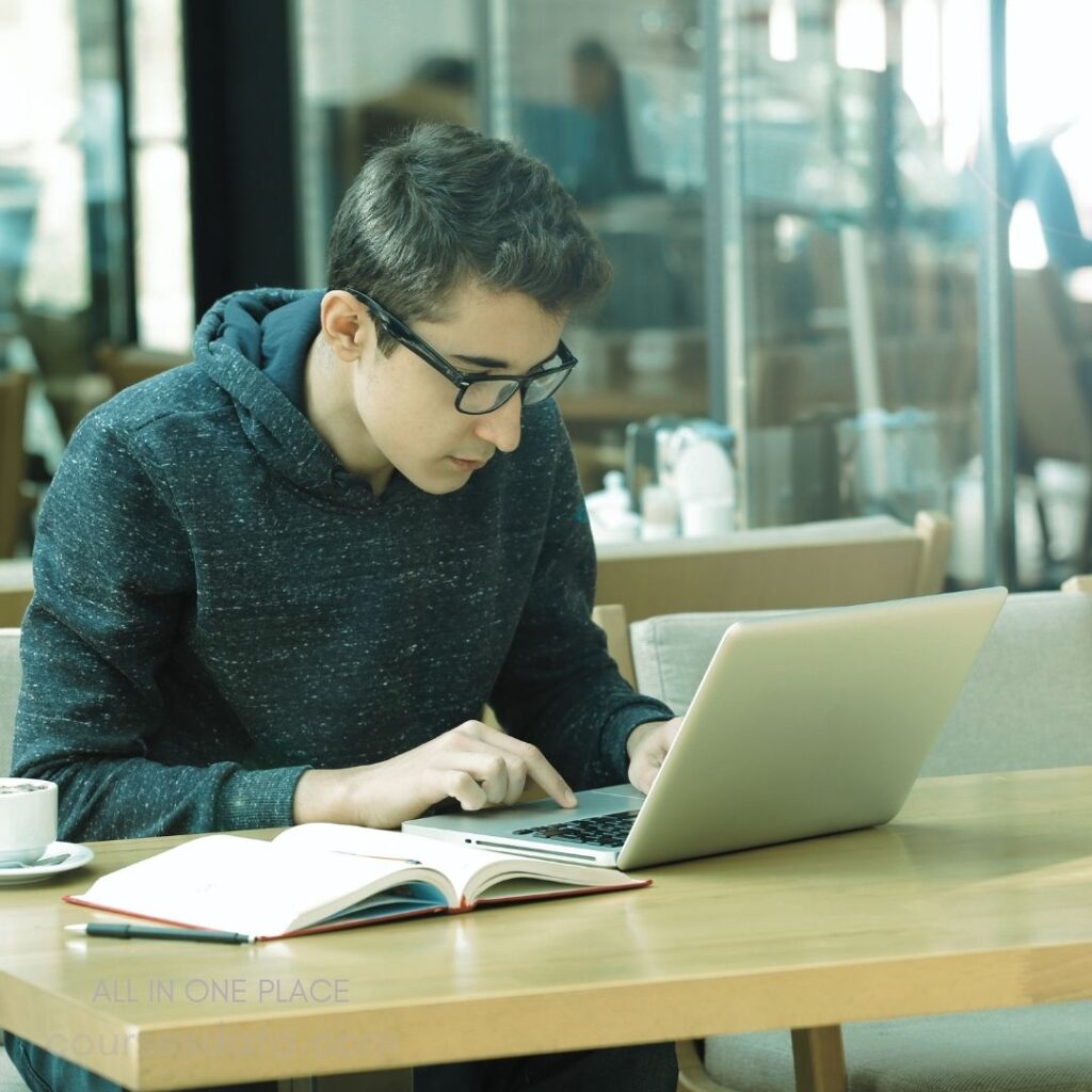 Young man working on laptop. Casual hoodie, focused expression. Open notebook and coffee cup. Bright, modern café setting. Natural light through windows.