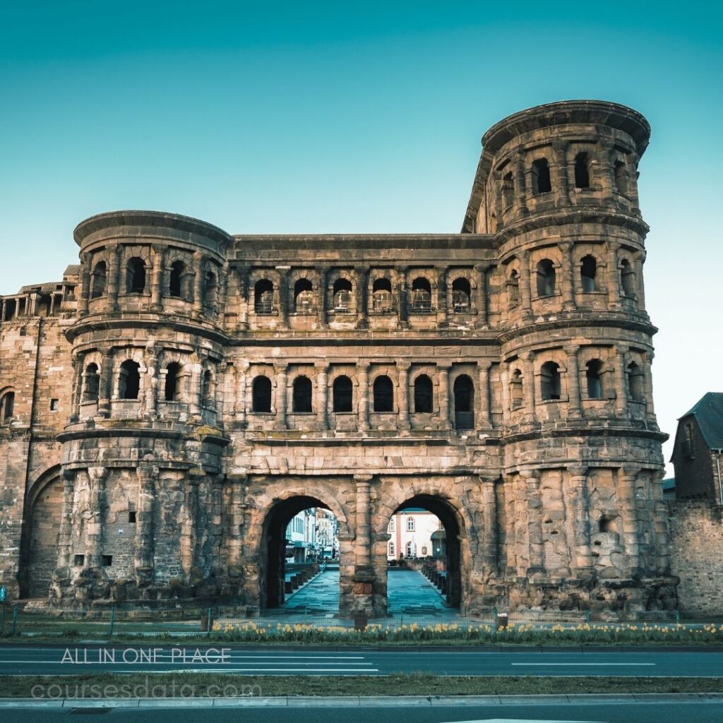 Ancient stone Roman structure. Multiple arched openings visible. Historical architecture under clear sky. Surrounding street and buildings present. Foreground greenery and flowers evident.