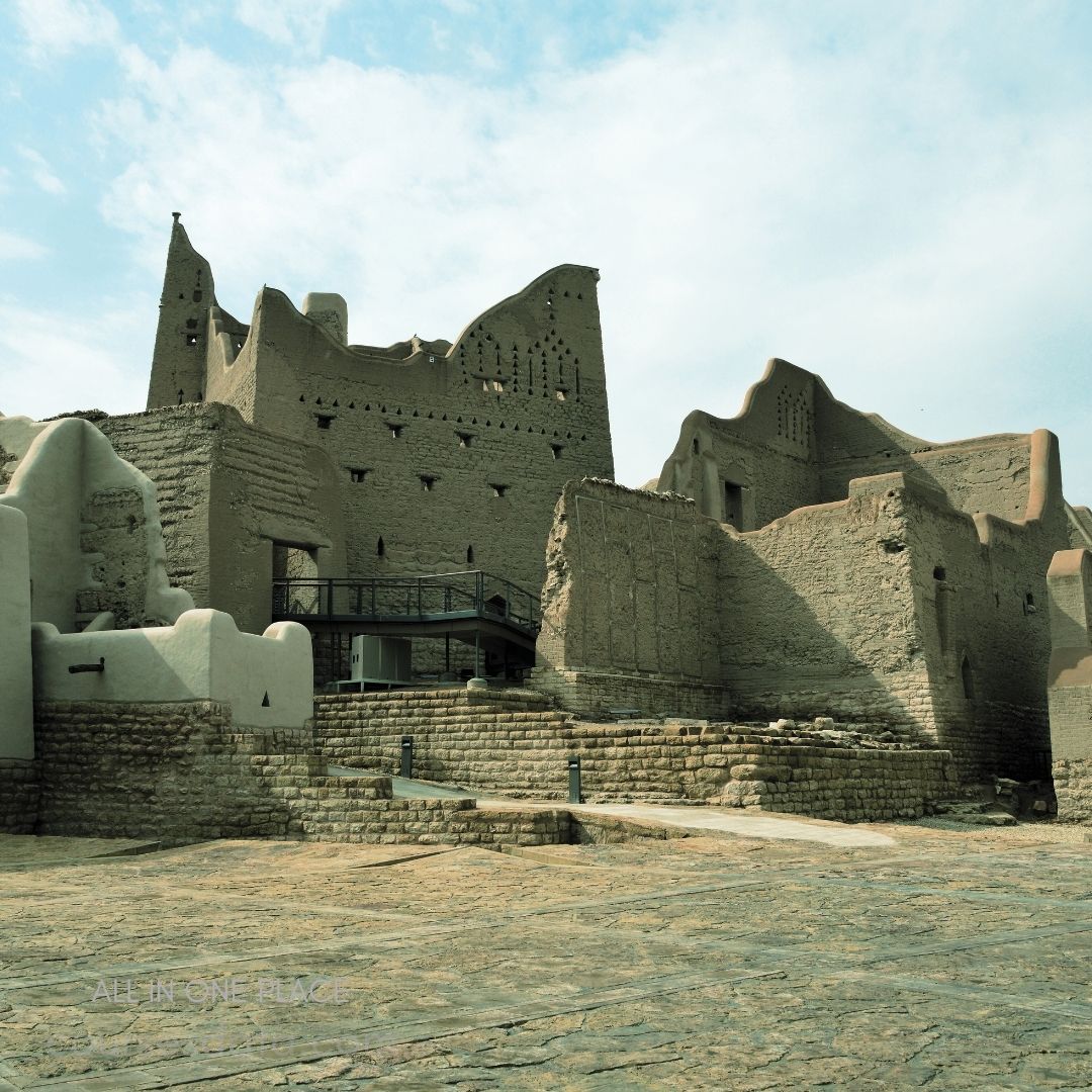 Historic mud-brick architecture. Stepped stone pathway. Sky with scattered clouds. Ancient ruins in foreground. Textured walls and patterns.