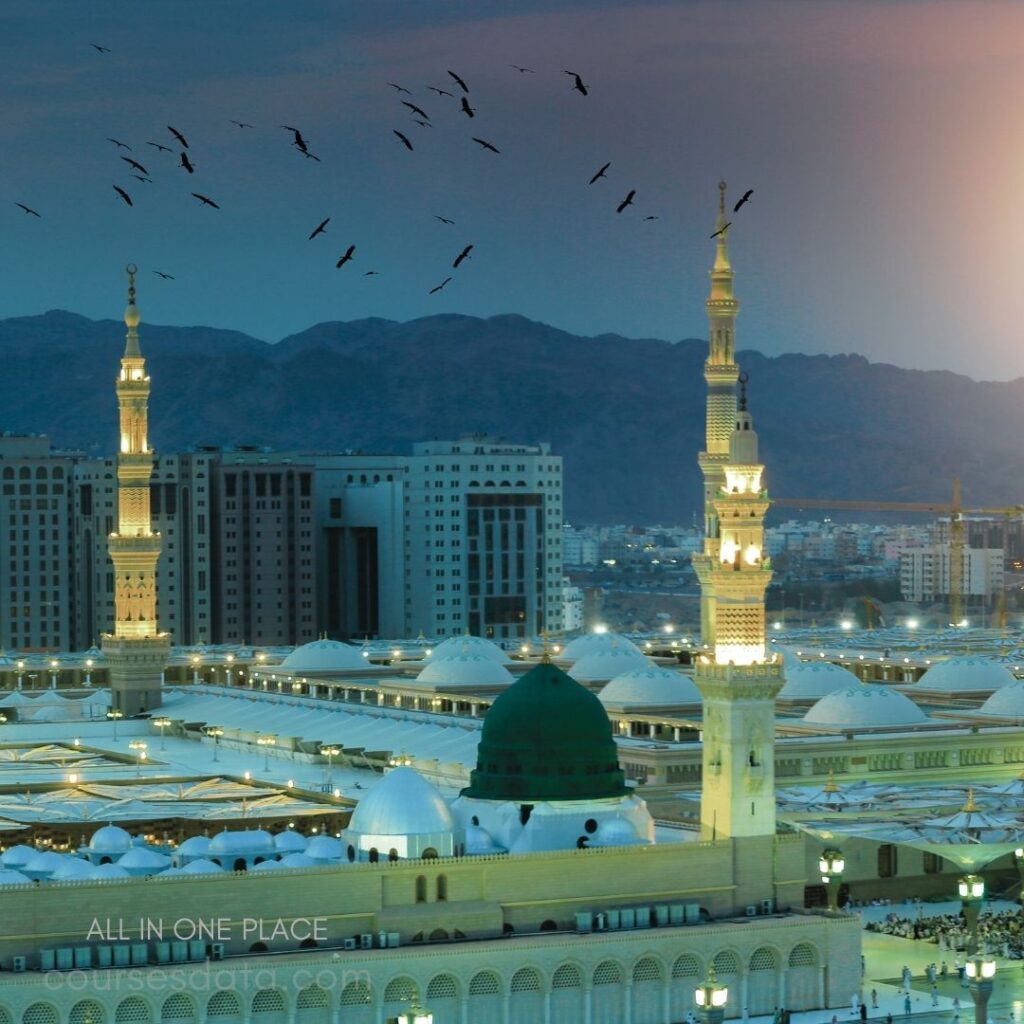 Mosque minarets at dusk. Green dome prominent. Birds flying above. City skyline background. Illuminated courtyard beneath.