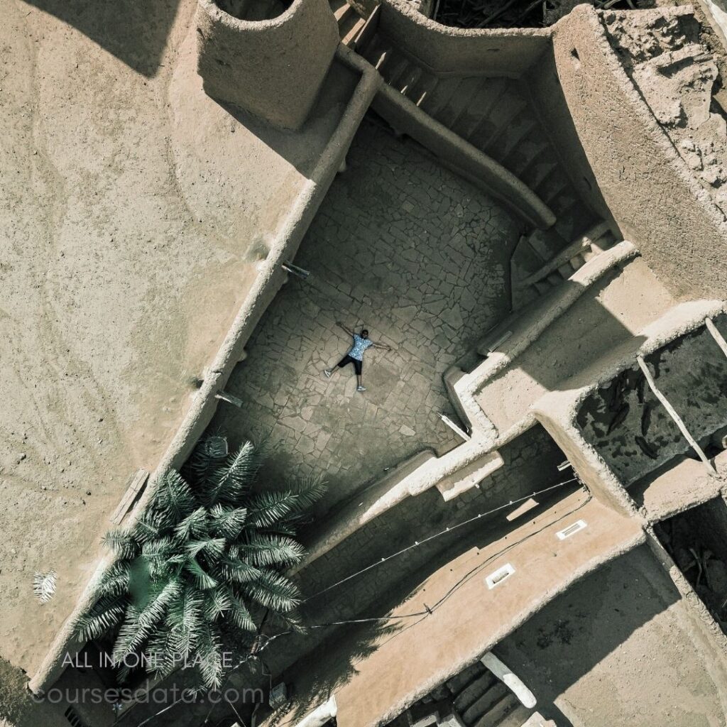 Aerial view of a person. Lying on stone courtyard. Surrounded by earthen walls. Nearby palm tree visible. Architectural ruins in background.