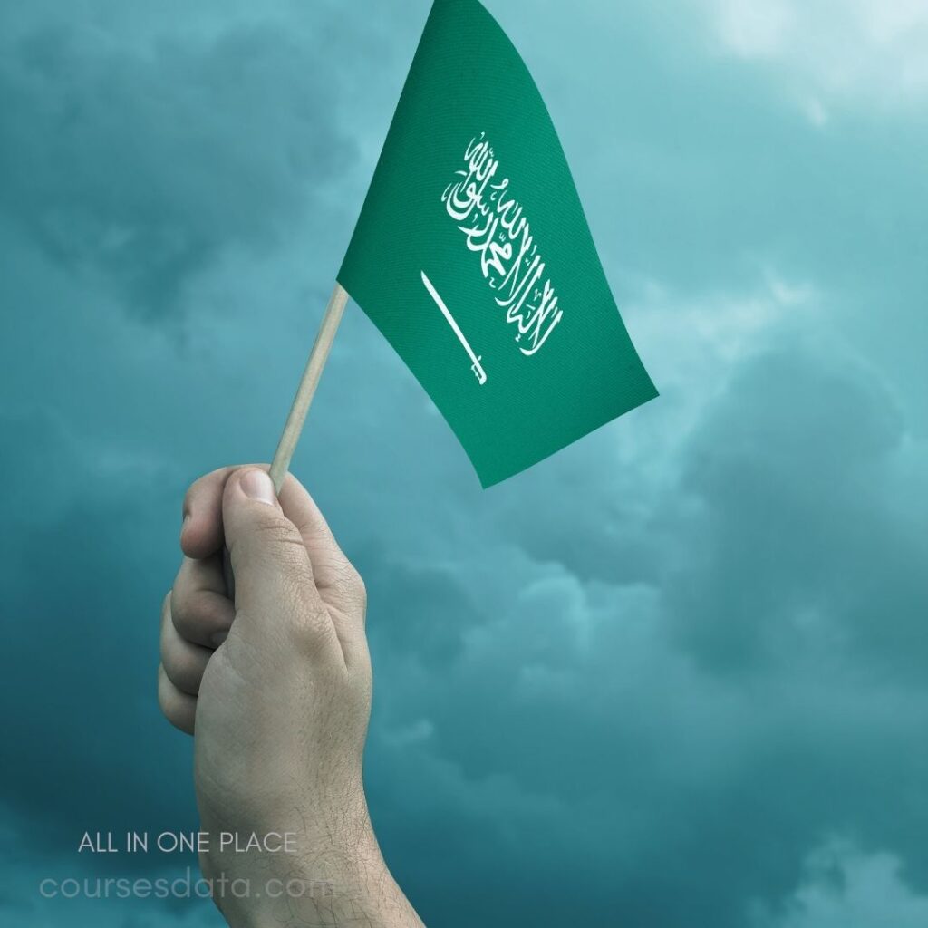 Hand holding Saudi flag. Green background. Cloudy sky. White Arabic text. Sword emblem visible.