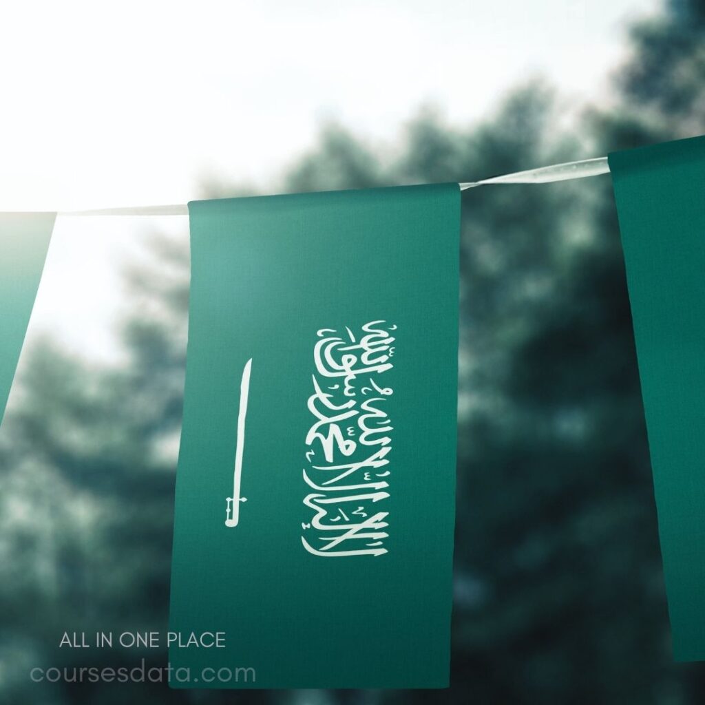 Green flag with Arabic text. White sword alongside the text. Background features blurred greenery. Sunlight illuminating the flag. Text signifies Saudi heritage.