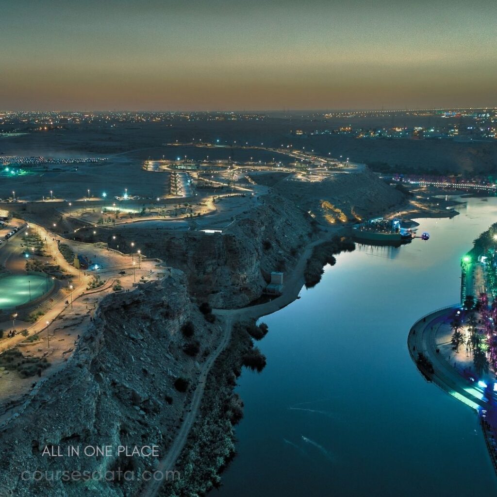 Scenic river view at dusk. Illuminated paths and greenery. Surrounding hills and city lights. Calm water reflecting evening sky. Colorful lights along riverbank.