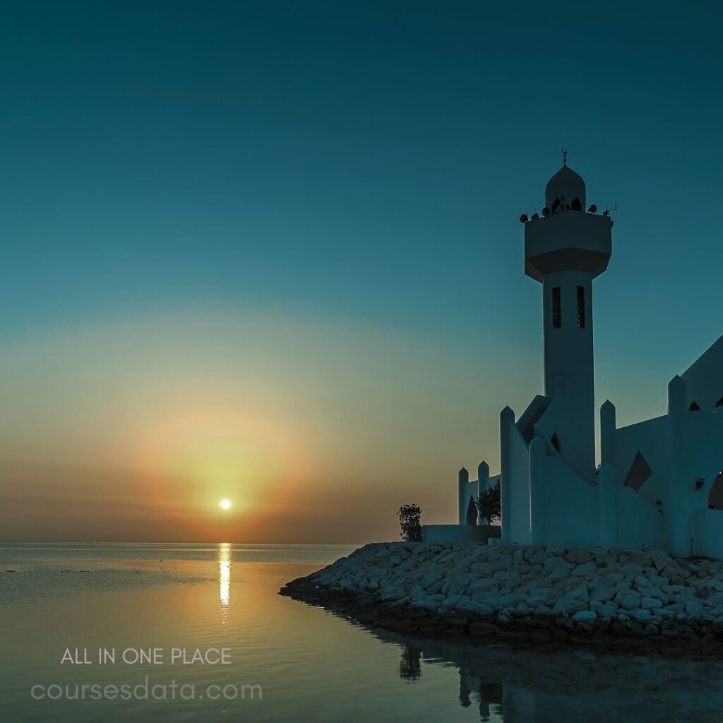 Sunset over calm waters. White mosque silhouette. Reflected lights on water. Blue sky gradient backdrop. Rocky shoreline with greenery.