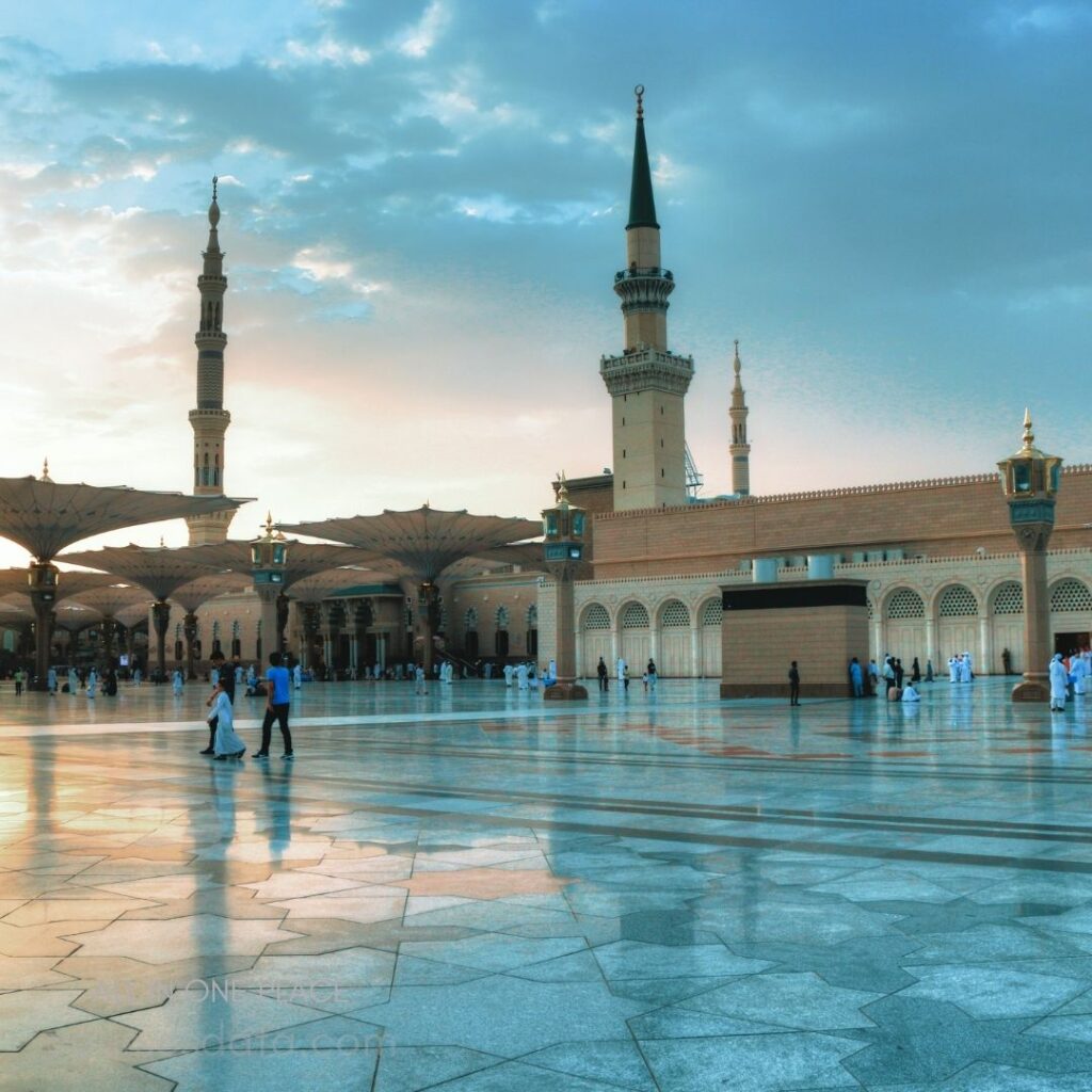 Mosque with tall minarets. Covered prayer area umbrellas. Reflective marble courtyard surface. People walking in prayer area. Evening sky with soft clouds.