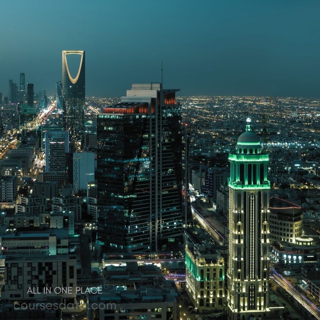 City skyline at dusk. Prominent skyscrapers illuminated. Distinctive ring-shaped building visible. Colorful lights along streets. Urban landscape with distant horizon.