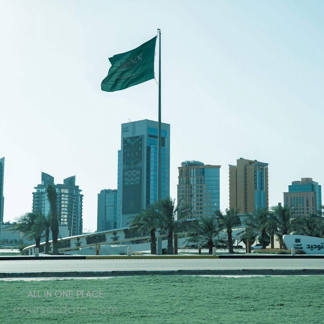 Saudi flag in urban setting. Modern buildings in background. Palm trees line the street. Clear sky, serene atmosphere. Cityscape of Saudi Arabia.