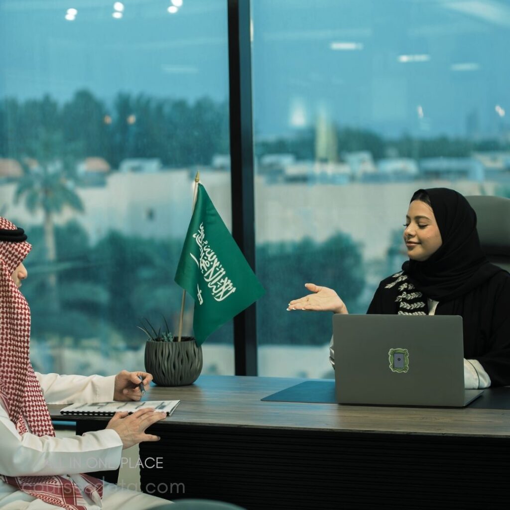 Business meeting, Saudi flag, professional attire, woman gesturing, man listening.