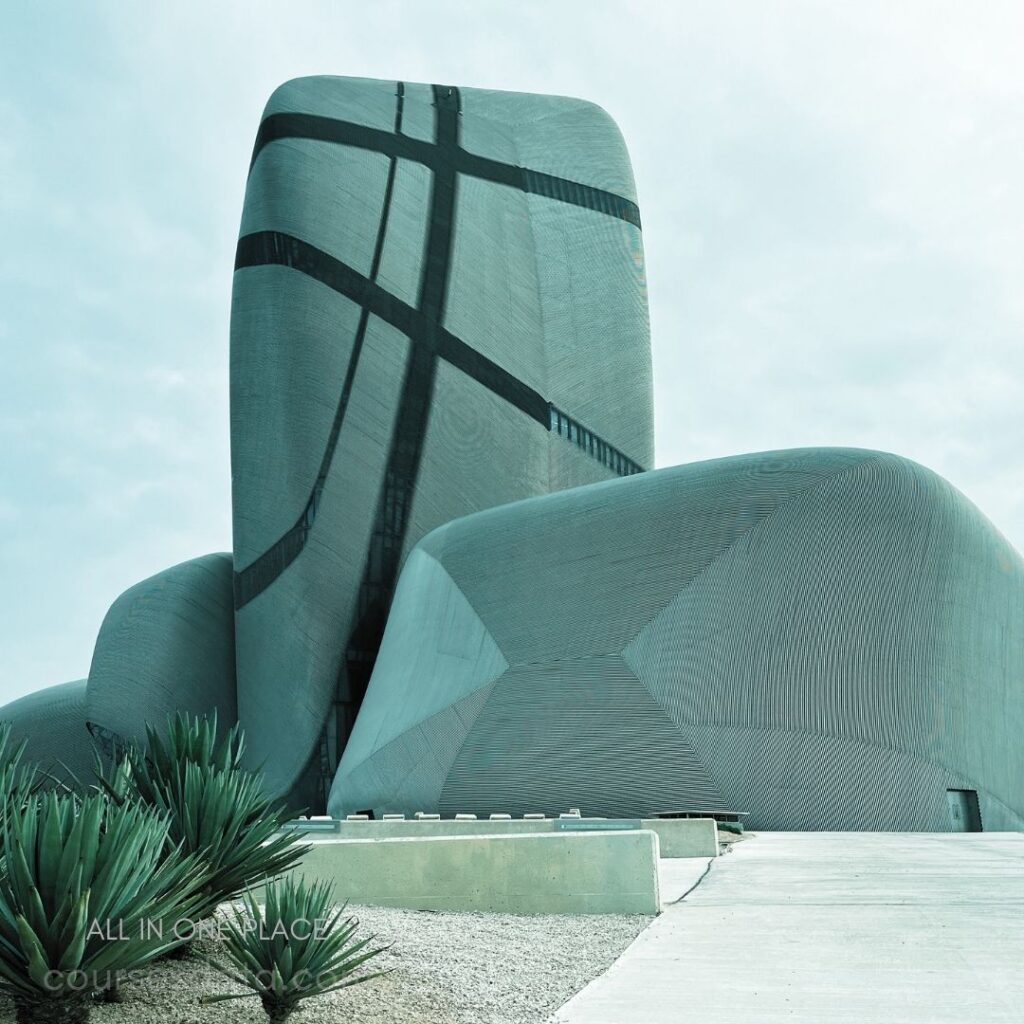 Modern sculptural architectural design. Curved surfaces with textured patterns. Surrounding desert landscaping features plants. Overcast sky creates muted tones.