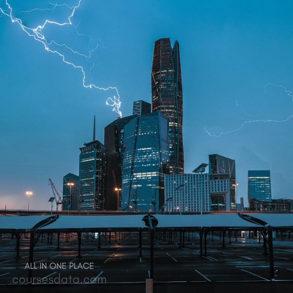 Lightning strikes modern skyscrapers. Dark blue sky backdrop. Empty parking area foreground. Illuminated building facades. Construction cranes visible nearby.