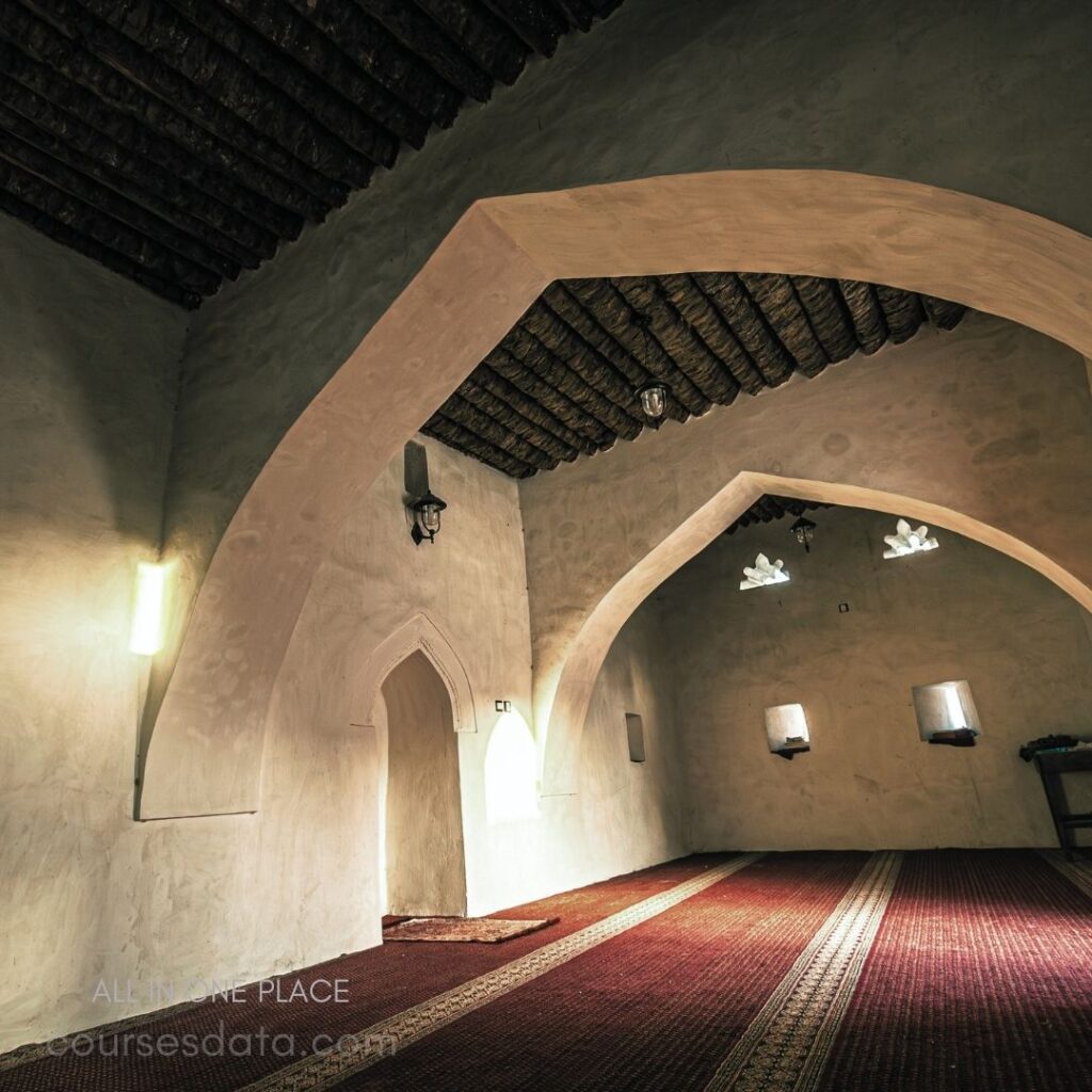 Interior of traditional mosque. Arched ceilings and walls. Decorative wooden beams above. Soft natural light filtering in. Colorful woven carpet covering floor.