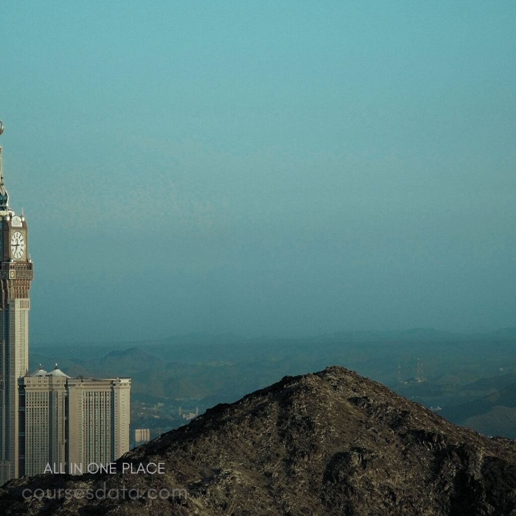 Skyline with tower structure. Mountain in foreground. Clear blue sky backdrop. Distant hills visible. Urban environment below.