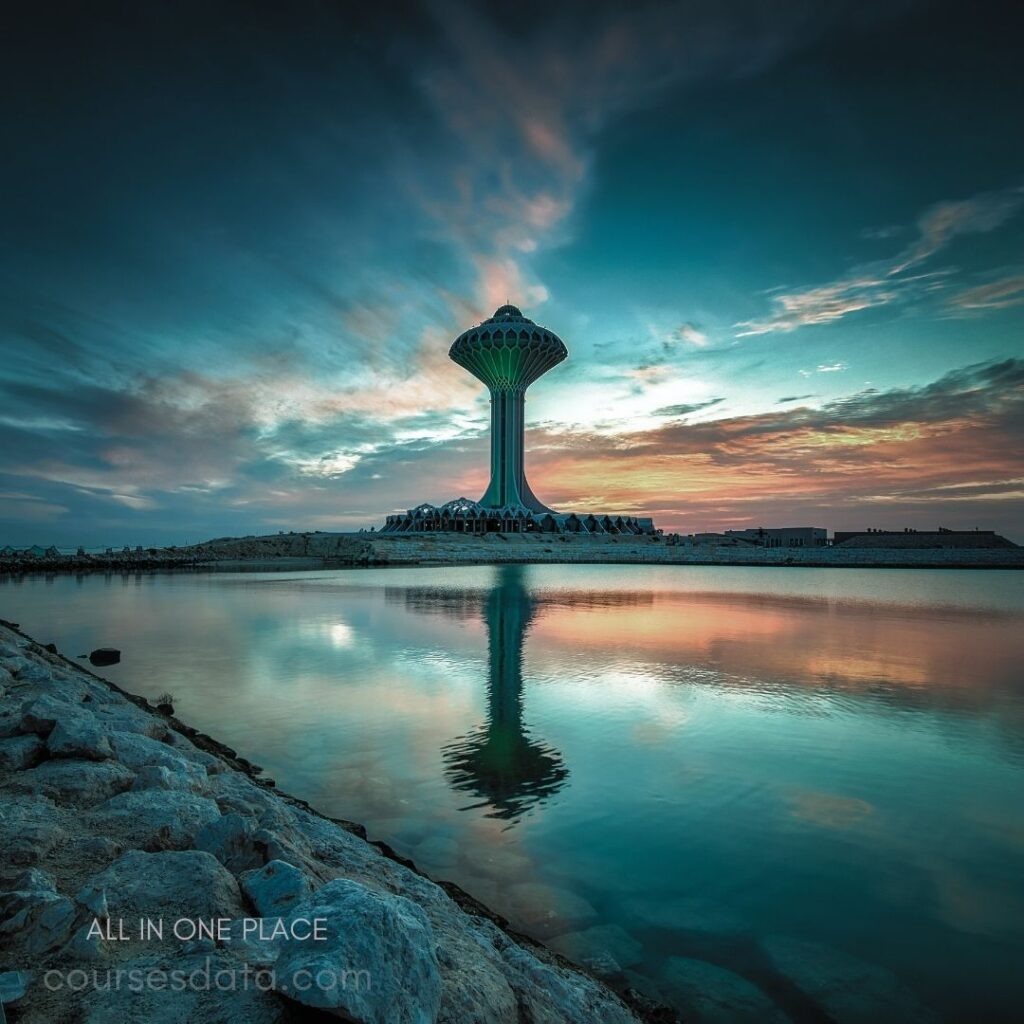 Tall tower with green lights. Calm water reflecting structure. Colorful sunset sky backdrop. Rocky shoreline in foreground. Serene and picturesque scene.