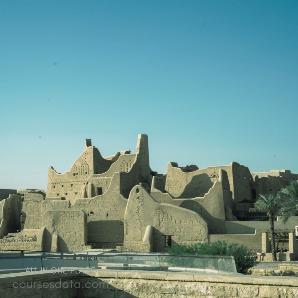 Mud brick structures with towers. Clear blue sky backdrop. Ancient architectural design features. Lush greenery in foreground. Desert landscape setting.