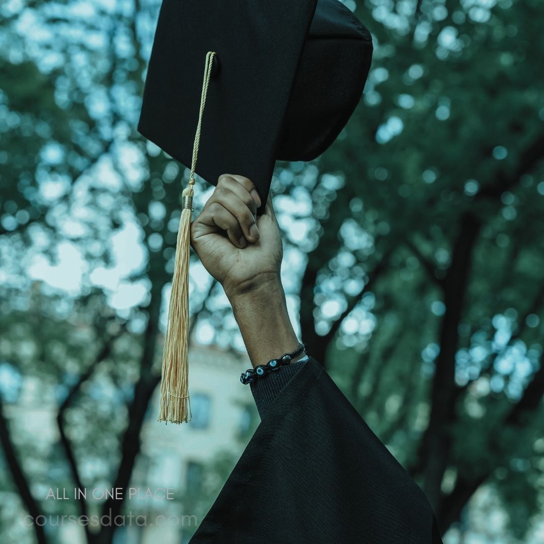 Graduation cap raised proudly. Hand wearing a bracelet. Blurred trees in background.
