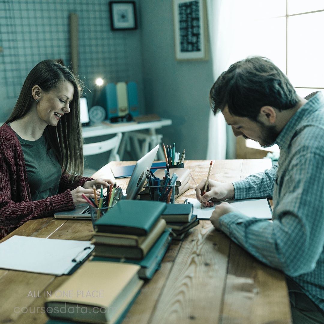 Two people studying together. Wooden table with notebooks. Colorful pens in holders. Laptop on the left. Natural light from window.
