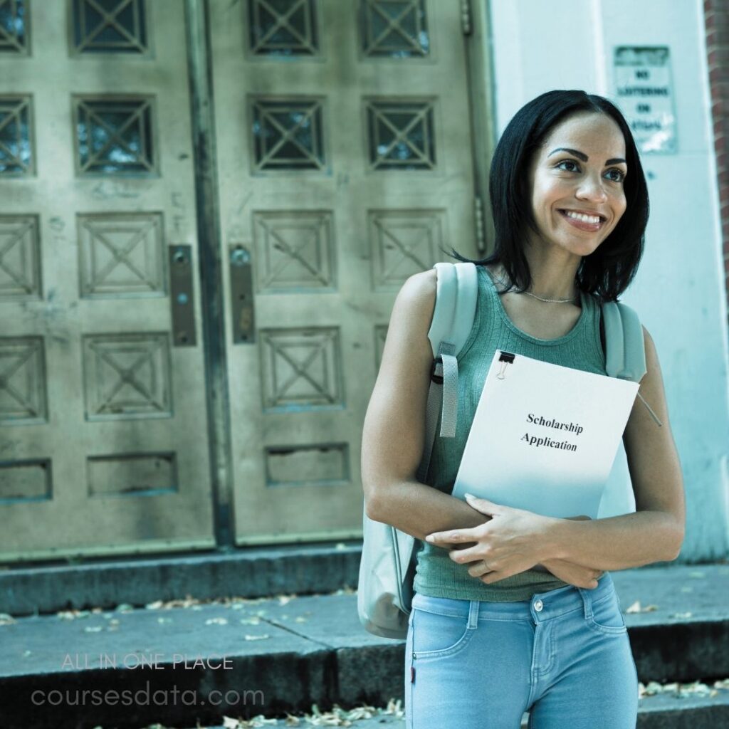 Smiling woman holding application. Wearing gray tank top. Backpack slung over shoulder. Golden doors in background. Outdoor setting with leaves.