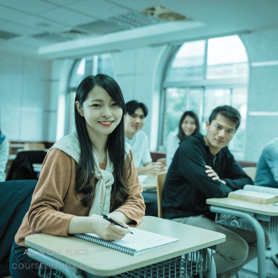 Smiling student in classroom. Notebook and pen in hand. Other students in background. Natural light through windows. Casual learning environment.
