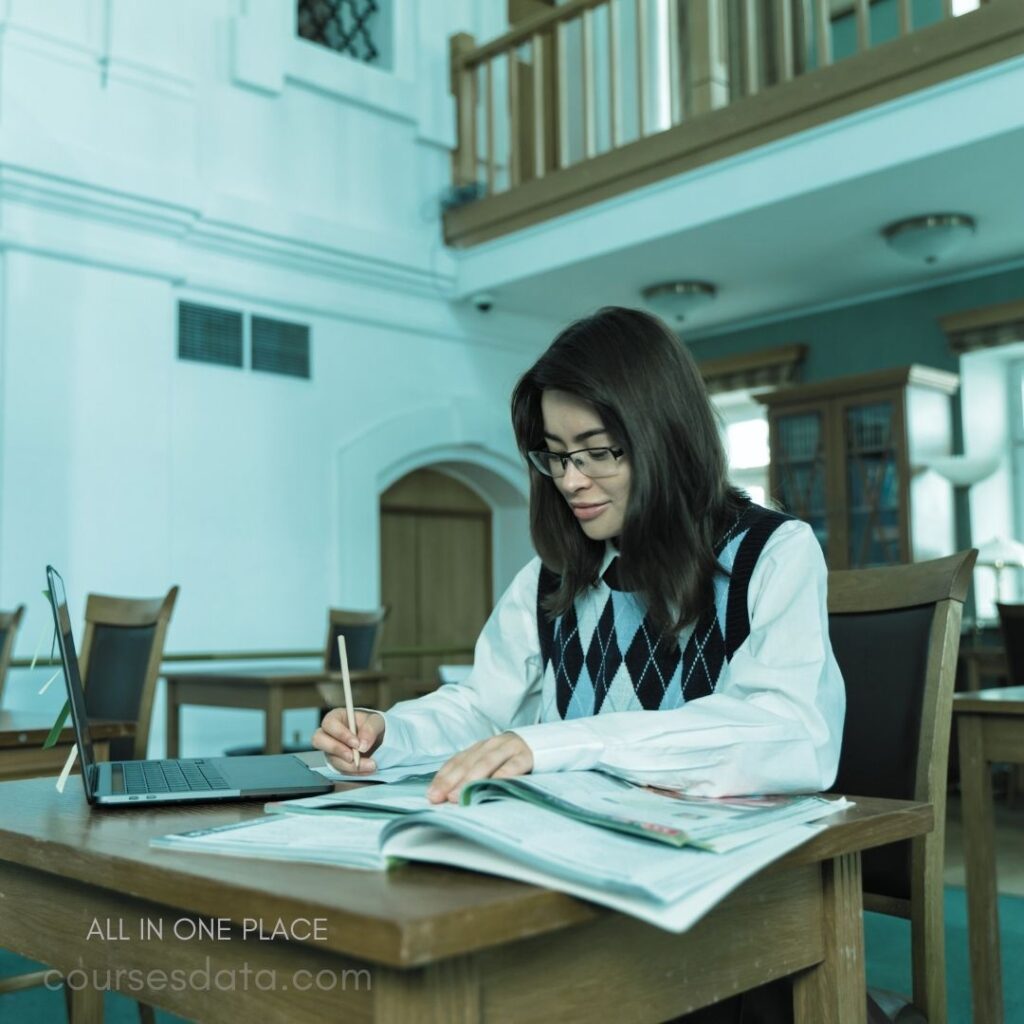 Student studying at desk. Notebook and laptop present. Library setting with large windows. Wearing glasses and a sweater. Focused expression, writing notes.