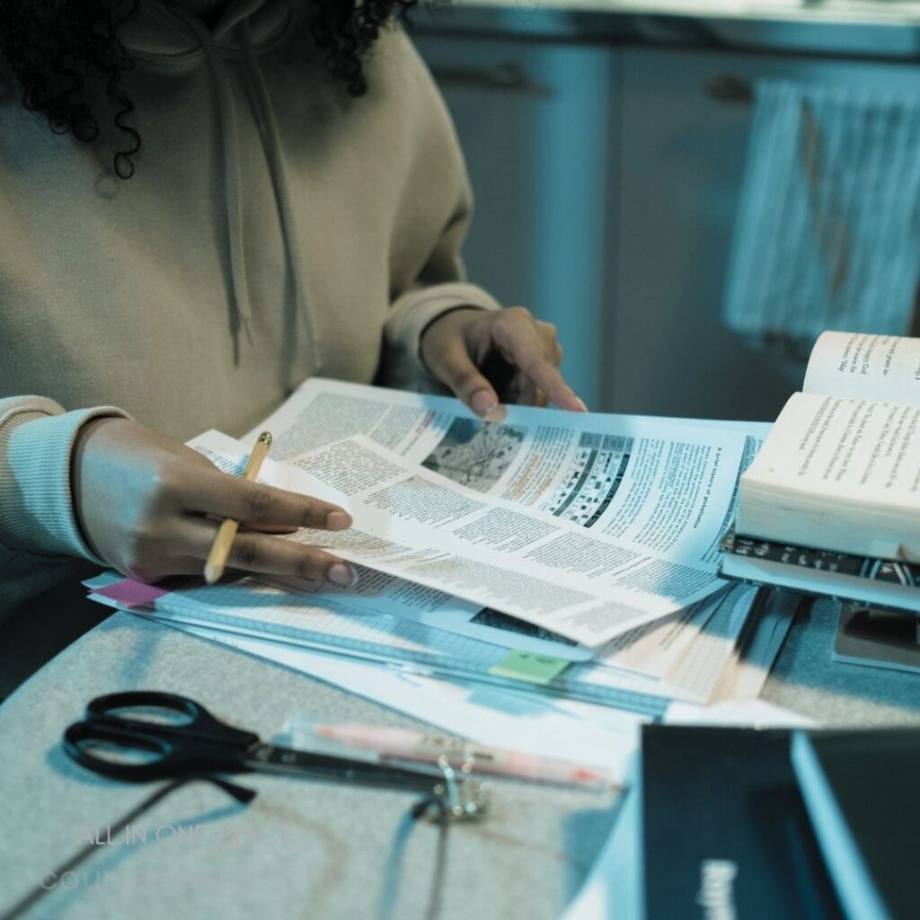 Person studying with papers. Hands holding documents. Scissors and pen nearby. Books stacked on table. Natural light from window.