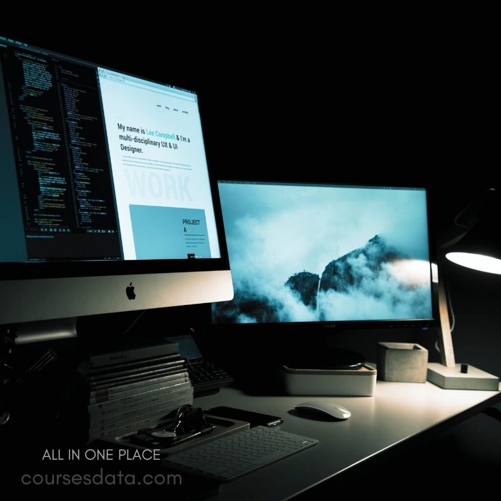 Two computer monitors, desk setup. Coding and design displayed. Lamp illuminates workspace. Stack of books nearby. Minimalist office environment.