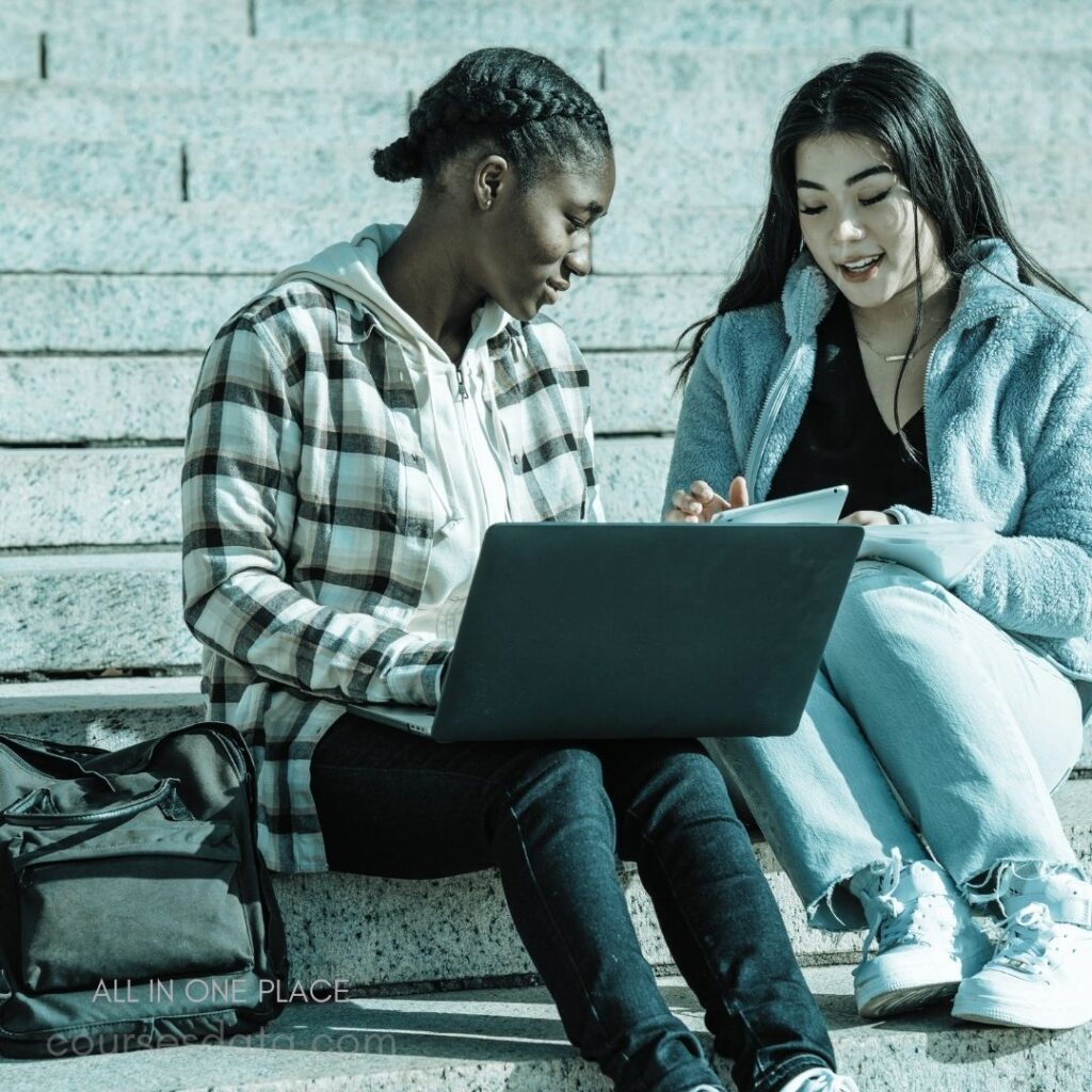 Two students studying together. Laptop and tablet on steps. Casual clothing and relaxed expressions. Diverse backgrounds engaged in discussion. Collaborative learning environment outdoors.