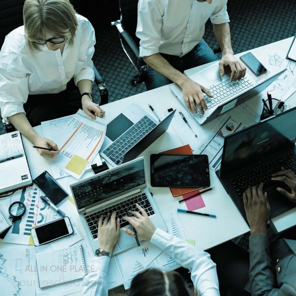 Group working at a table. Multiple laptops and papers. Hands typing and writing. Office setting with documents. Colorful sticky notes scattered.