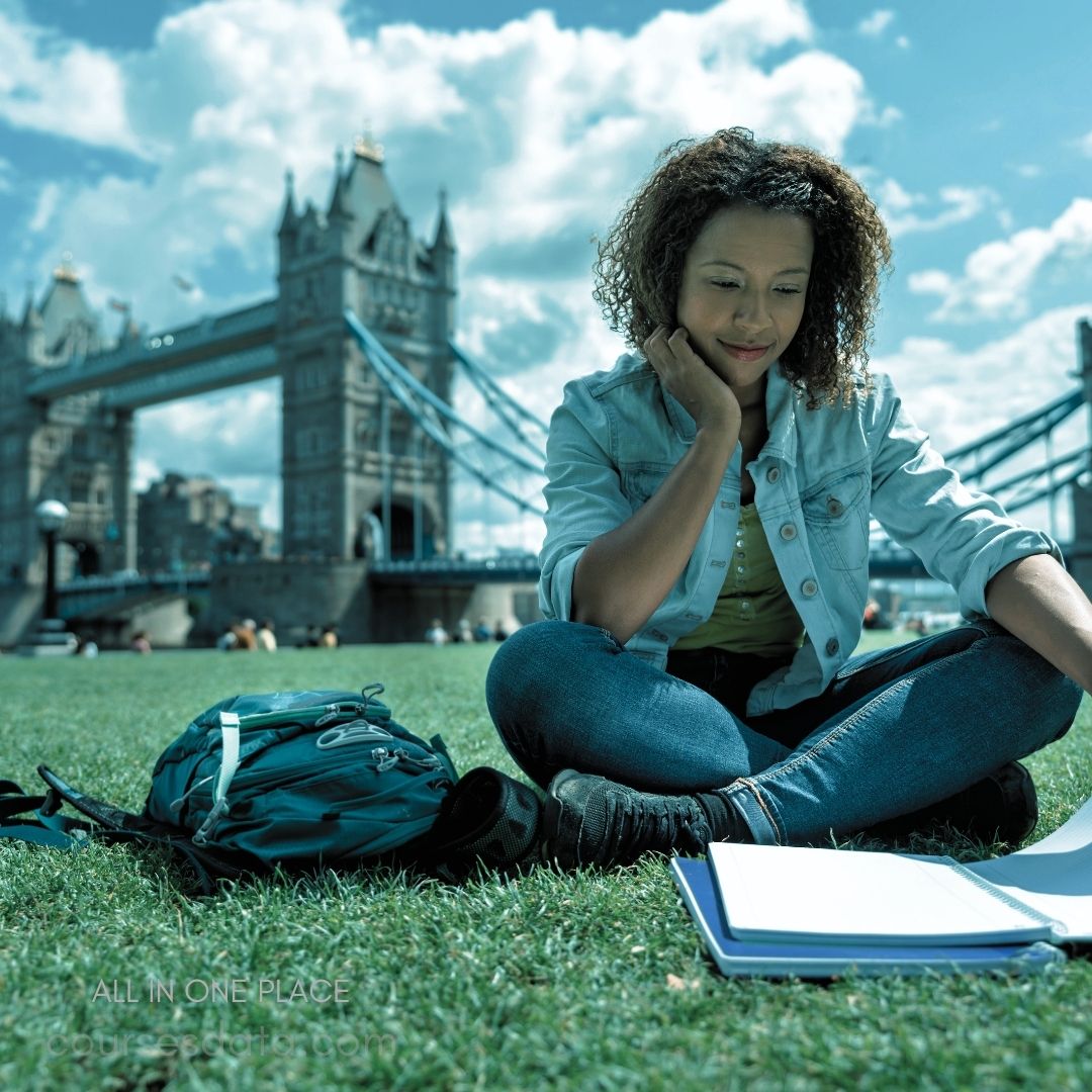 Young woman studying outdoors. Tower Bridge in background. Green grass, sunny sky. Backpack beside her. Open notebook on ground.
