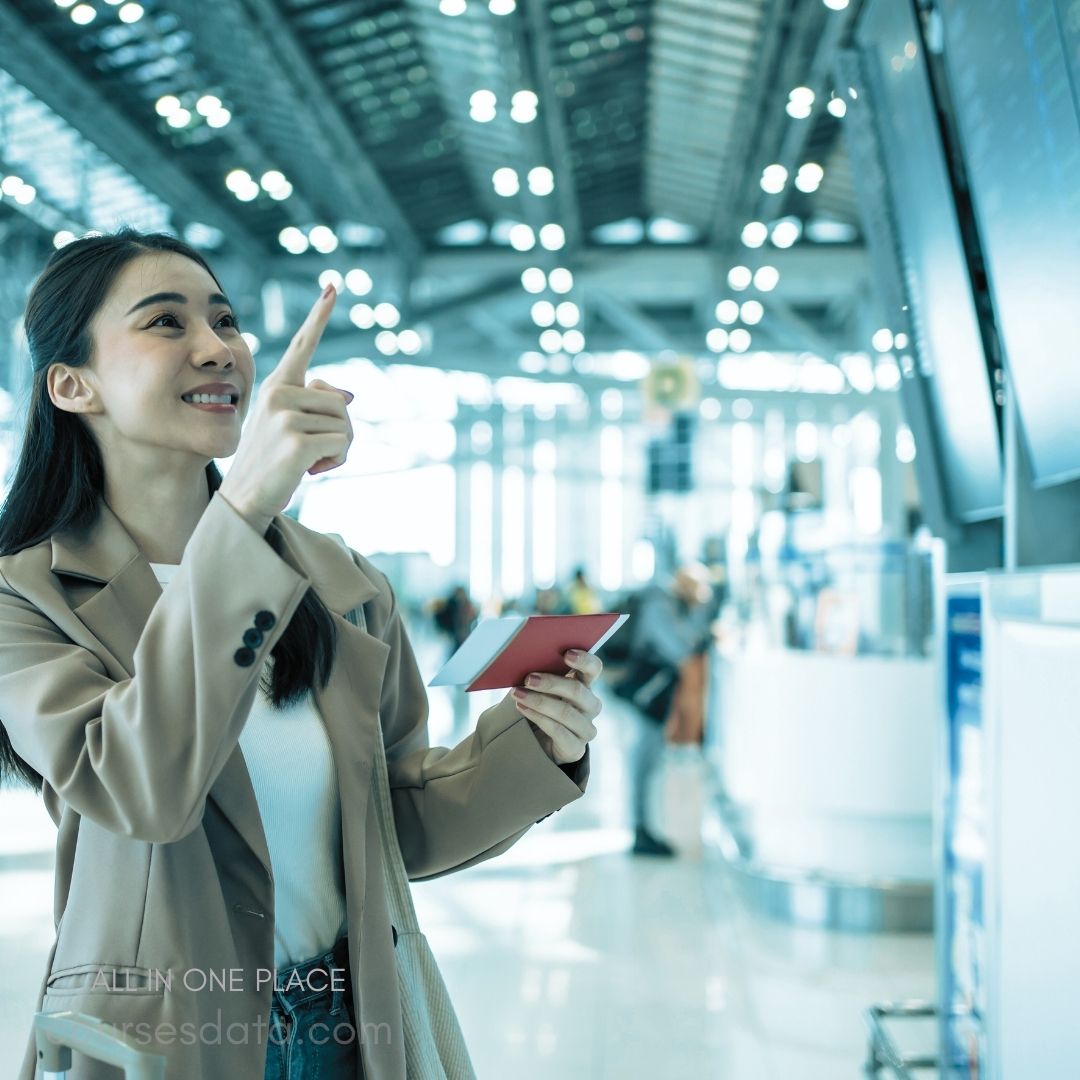 Woman at airport terminal. Holding red travel document. Pointing at information screen. Bright, modern architecture. Busy terminal atmosphere.