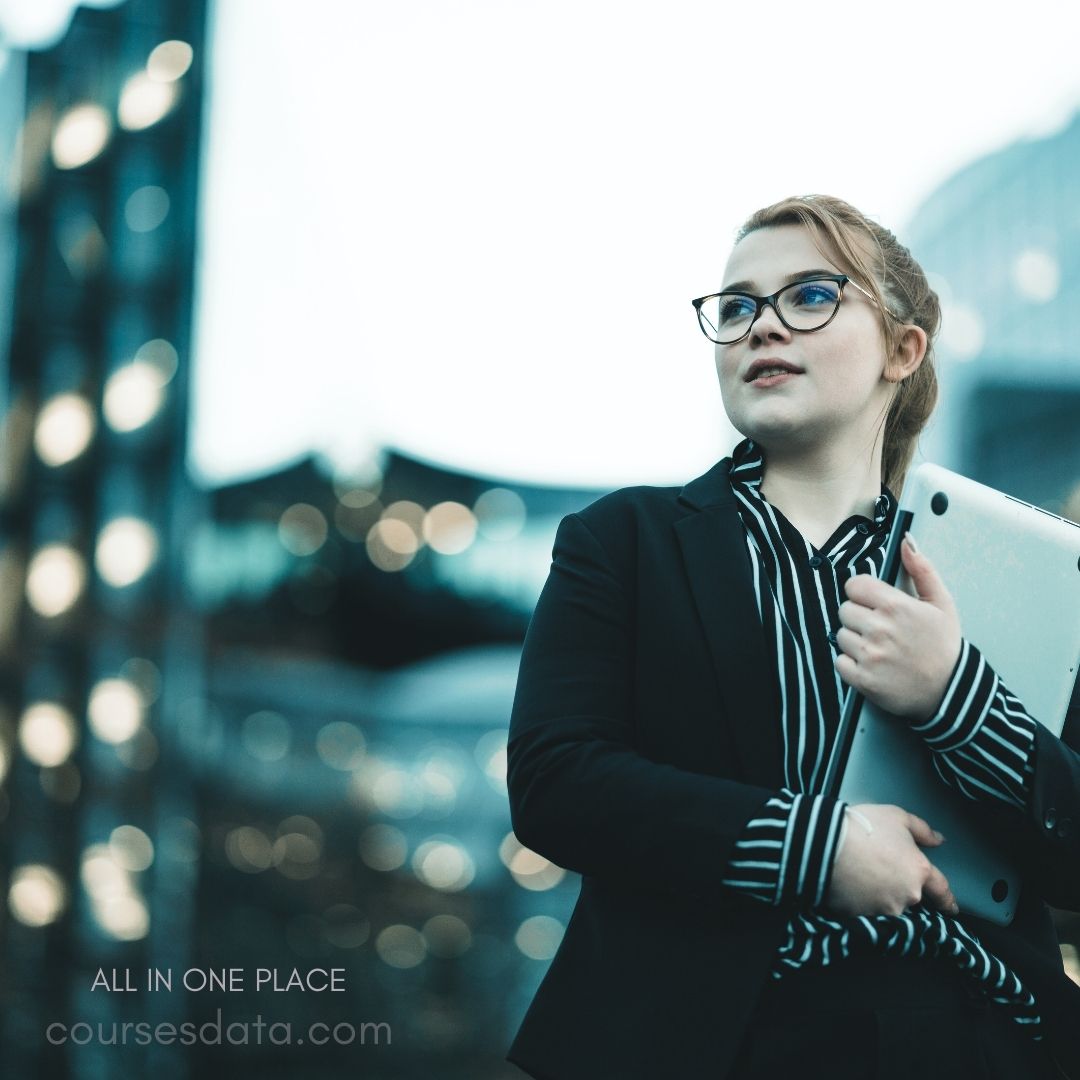 Businesswoman in modern setting. Holding a laptop. Wearing glasses and blazer. Confident expression outdoors. Urban background with lights.