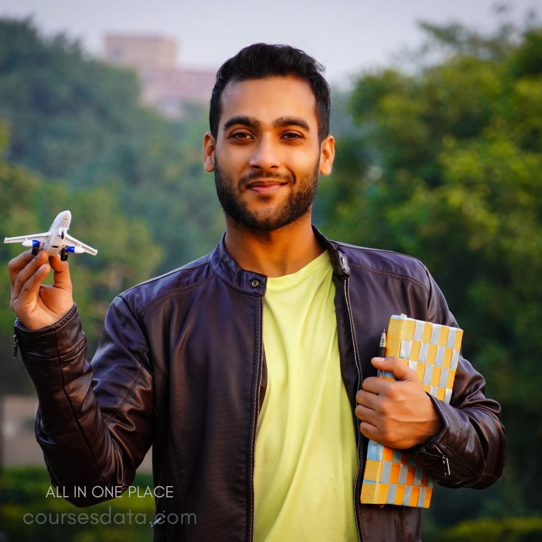 Smiling man holding toy airplane. Wearing leather jacket and yellow shirt. Carrying notebook in other hand. Green background suggests outdoor setting.