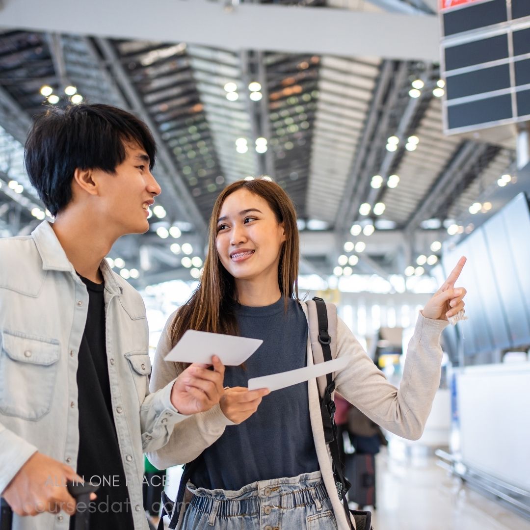 Two young travelers smile, engaging. Airport background shines brightly. They hold travel documents. One points, appearing inquisitive.