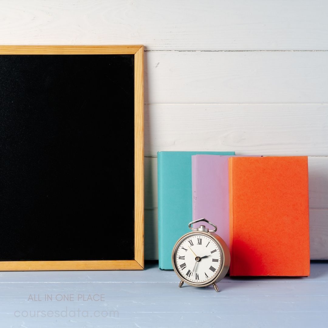 Blackboard with wooden frame. Three colorful books stacked. Silver alarm clock in front. Light wooden background texture. Blue surface beneath objects.